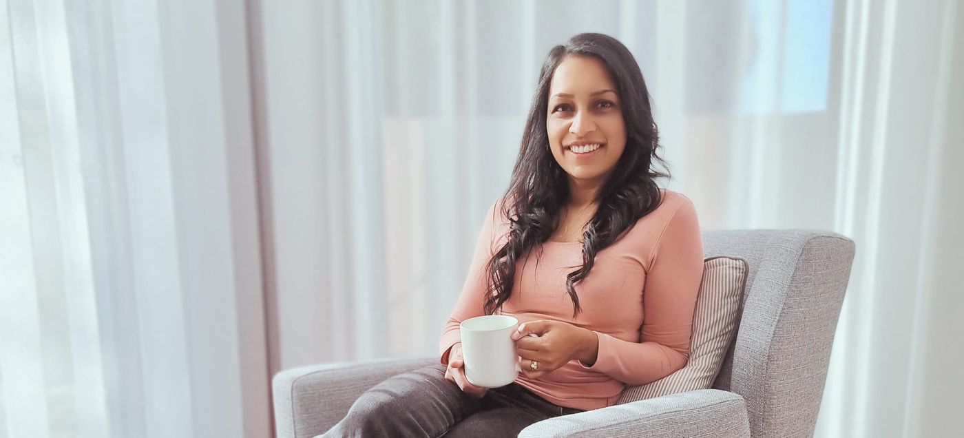 Nicole Jayawickreme sitting on a sofa by a window with a cup of tea, smiling