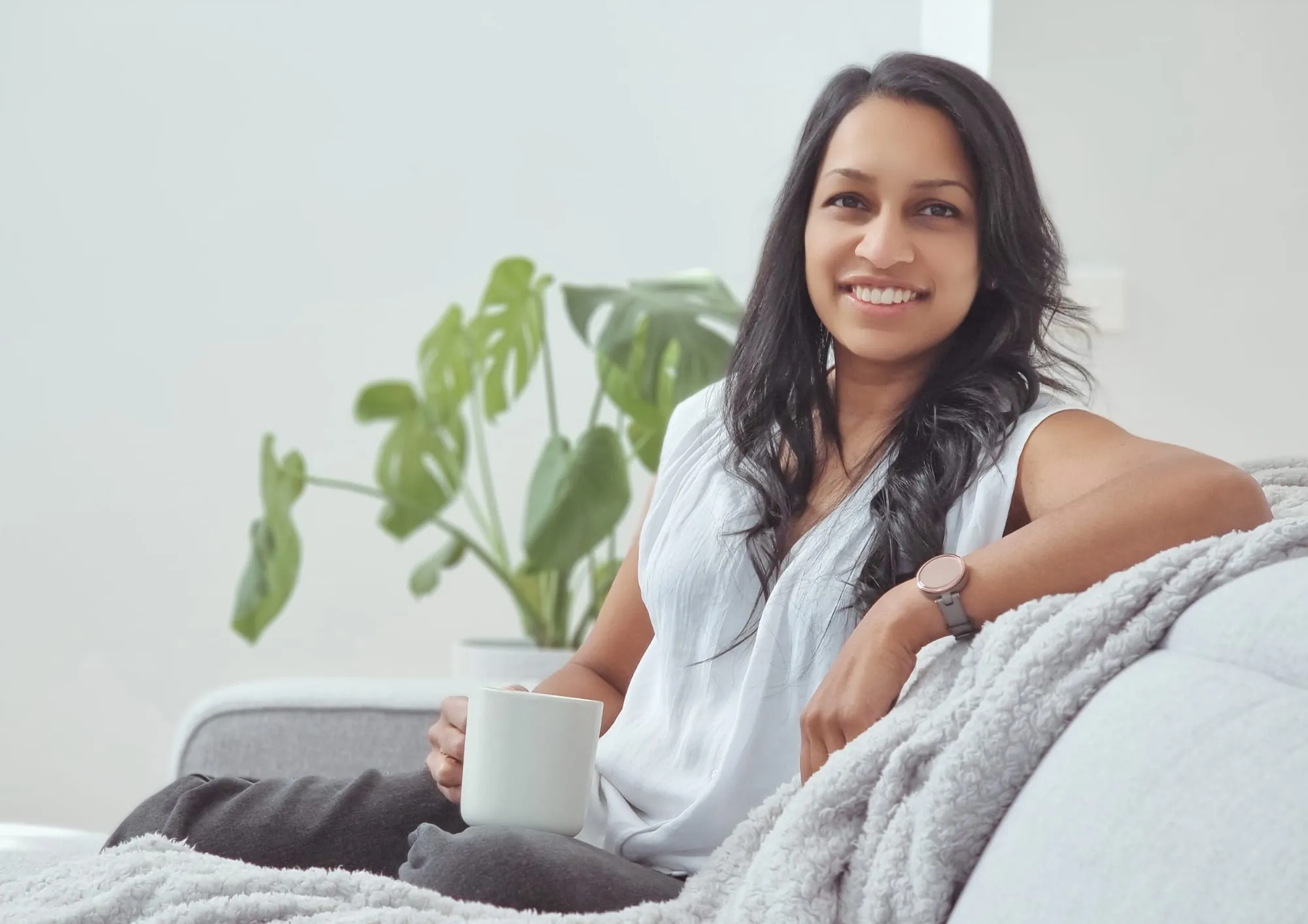 Nicole Jayawickreme on a sofa by a window with a plant, holding a cup